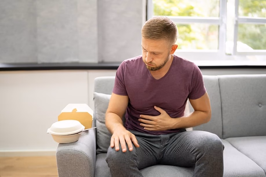 Man sitting on sofa holding his stomach in discomfort after eating takeaway food, illustrating digestive pain and heartburn symptoms