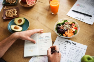 Person writing a nutrition plan at a wooden table with avocado, nuts, pomegranate, a salad bowl and dietary reference charts